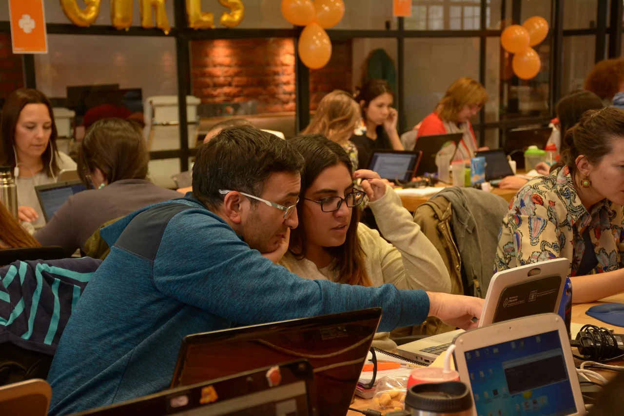 Man and woman working together on a laptop in a busy, collaborative classroom setting filled with students using computers.